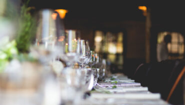 A blurred image of an elegantly set dining table at an Indian restaurant in Elstree. Glassware, including wine glasses and water glasses, are neatly arranged alongside white plates and cutlery. Soft lighting creates a warm ambiance, with greenery visible on the table in the foreground. | Indian Restaurant Elstree Borehamwood