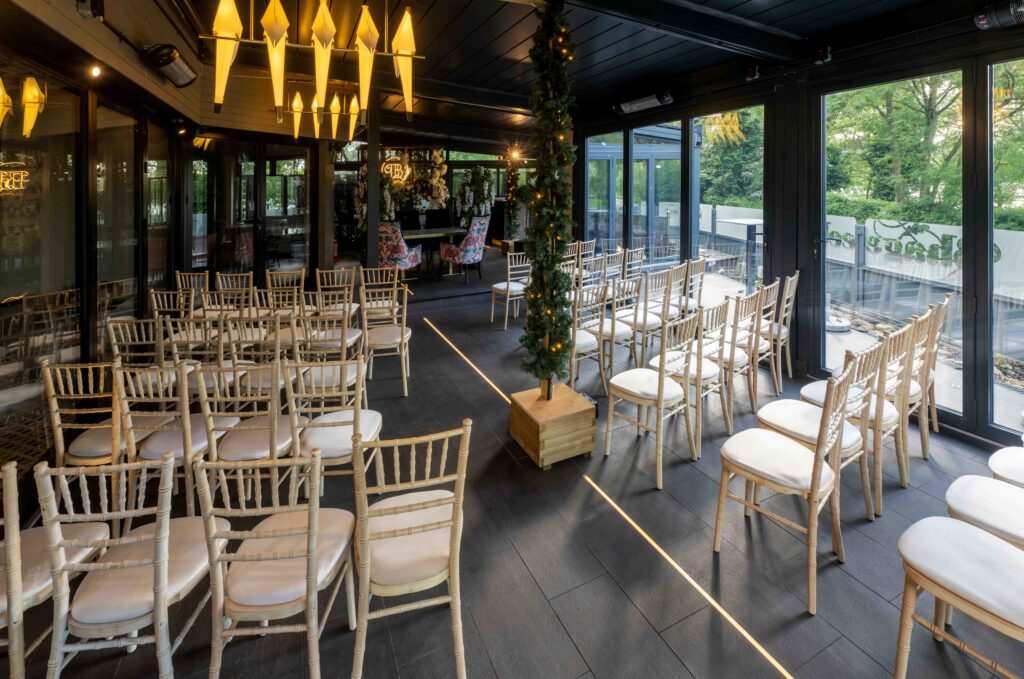 The image captures a serene space reminiscent of an Indian Restaurant in Borehamwood, with rows of wooden chairs facing an altar. Large windows reveal greenery, while modern yellow pendant lights and a decorative tree in the aisle complement the warm dark wood flooring. | Indian Restaurant Elstree Borehamwood