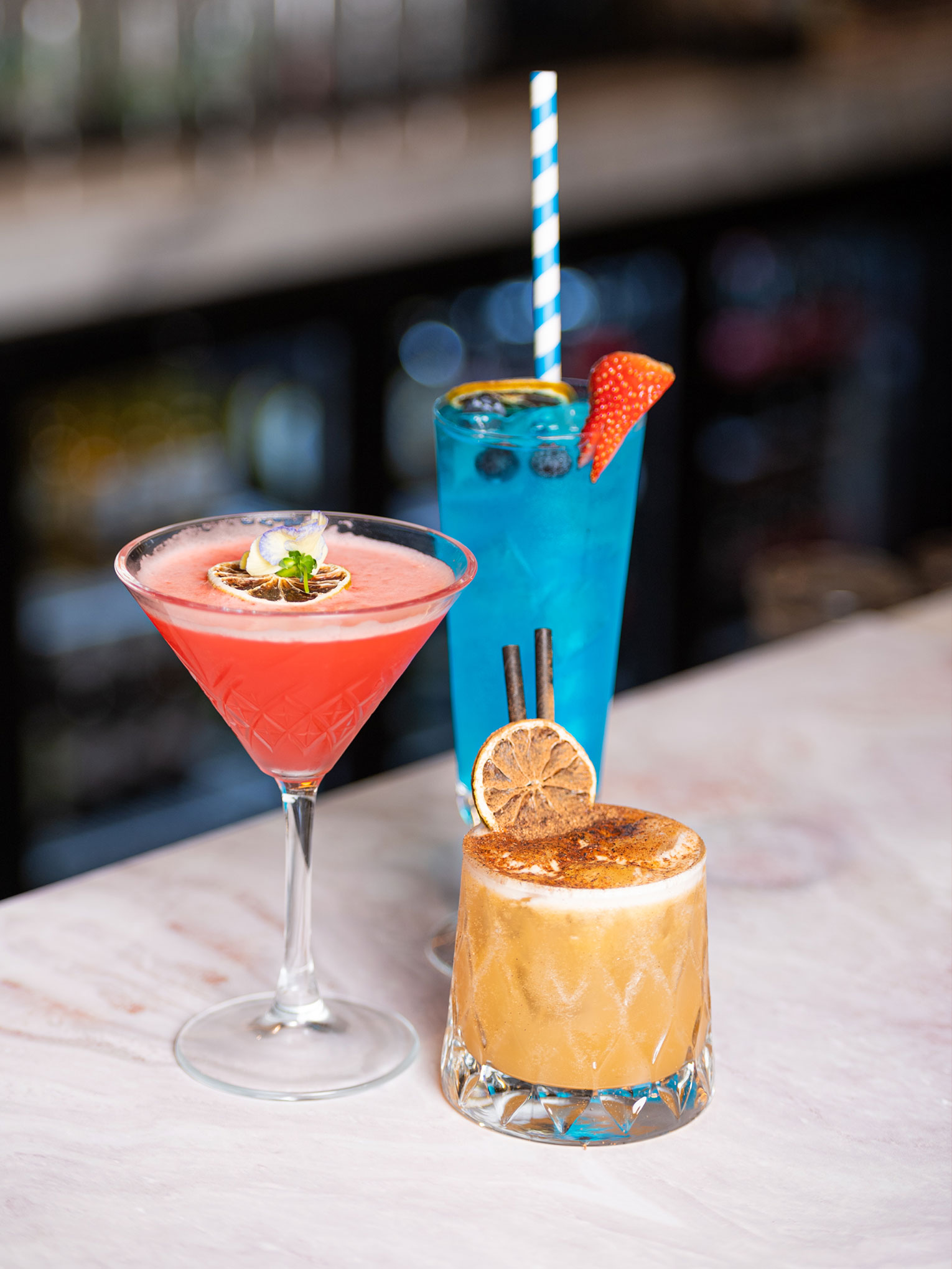 Three cocktails on a bar at an Indian Restaurant in Stanmore: a red martini with lime garnish, a tall blue drink with blueberries and strawberry, and a brown cocktail with dried orange slices and cinnamon stick. Each is served in distinct glassware on a marble surface. | Indian Restaurant Elstree Borehamwood