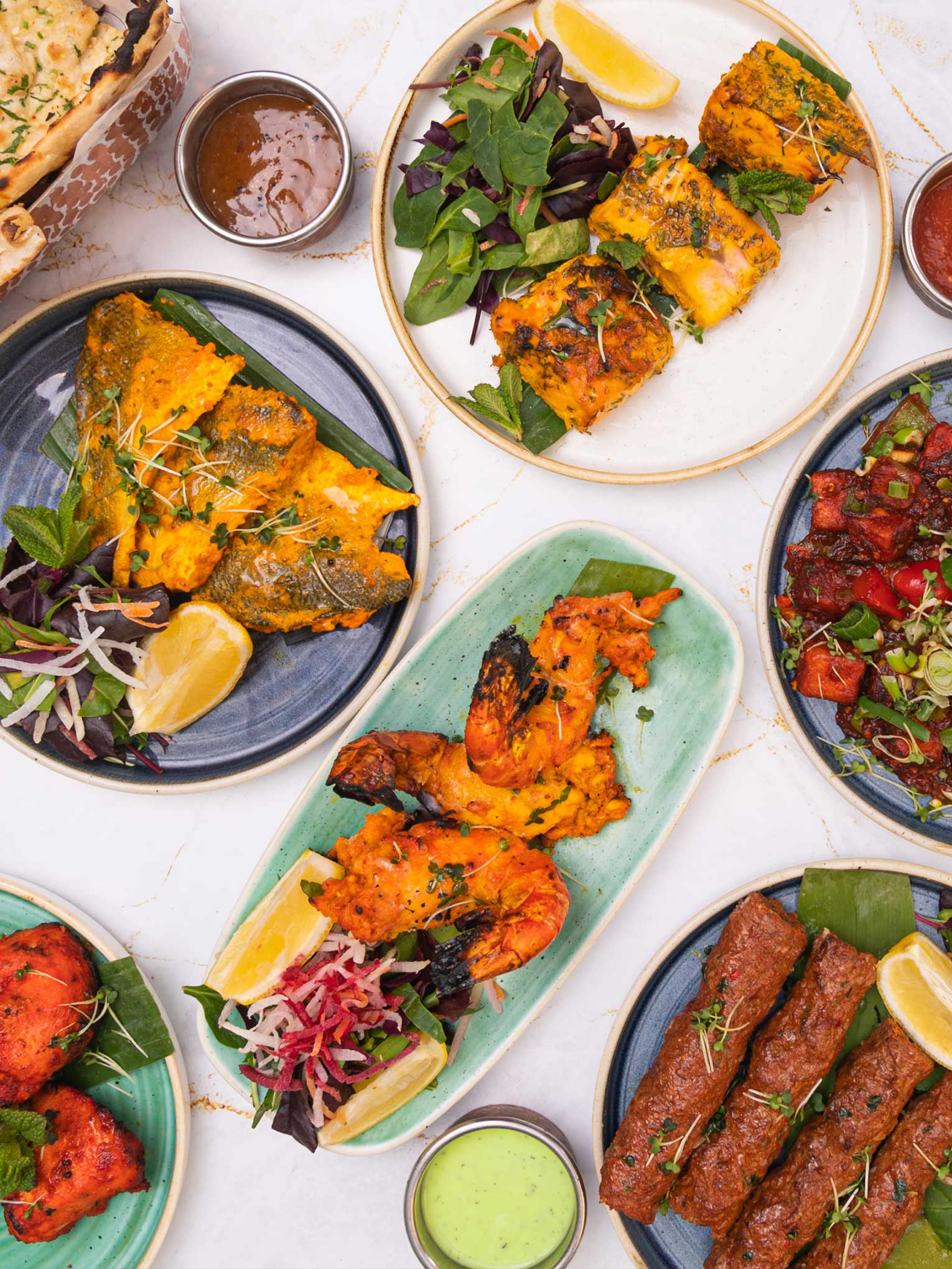 A variety of Indian dishes from an Indian takeaway in Elstree are displayed on a white marble surface. Plates include grilled shrimp, fish, chicken tikka, kebabs, and fresh greens, garnished with lemon wedges. Accompanying sauces are in small metal bowls, and a basket of naan is visible in the corner. | Indian Restaurant Elstree Borehamwood