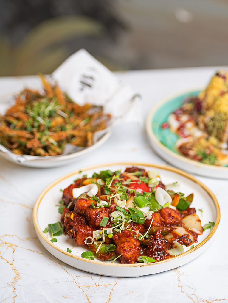 A plate of spicy glazed chicken topped with green onions and sprouts sits invitingly in the foreground. Behind it, a bowl of crispy fried vegetable strips and a dish with corn slightly blur into the background, all beautifully arranged on a white marble table at an Indian Restaurant in Elstree. | Indian Restaurant Elstree Borehamwood