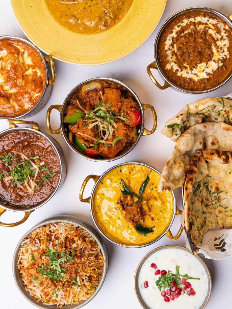 A variety of Indian dishes are arranged on a white background. Visible are bowls of colorful curries, rice, naan bread, and raita. The vibrant sauces and garnishes illustrate a diverse meal presentation from an Indian restaurant in Stanmore. | Indian Restaurant Elstree Borehamwood