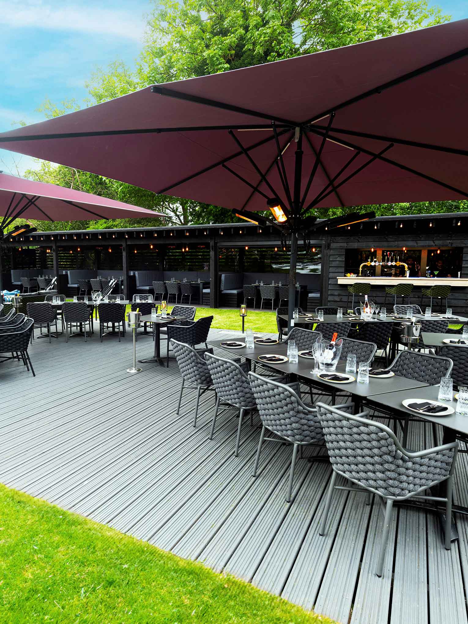 Experience alfresco dining at our Indian Restaurant Bushey. The outdoor area features dark wicker chairs and tables set for meals on a wooden deck, with large maroon umbrellas providing shade. A bar with stools and string lights complements the lush greenery under a partly cloudy sky. | Indian Restaurant Elstree Borehamwood