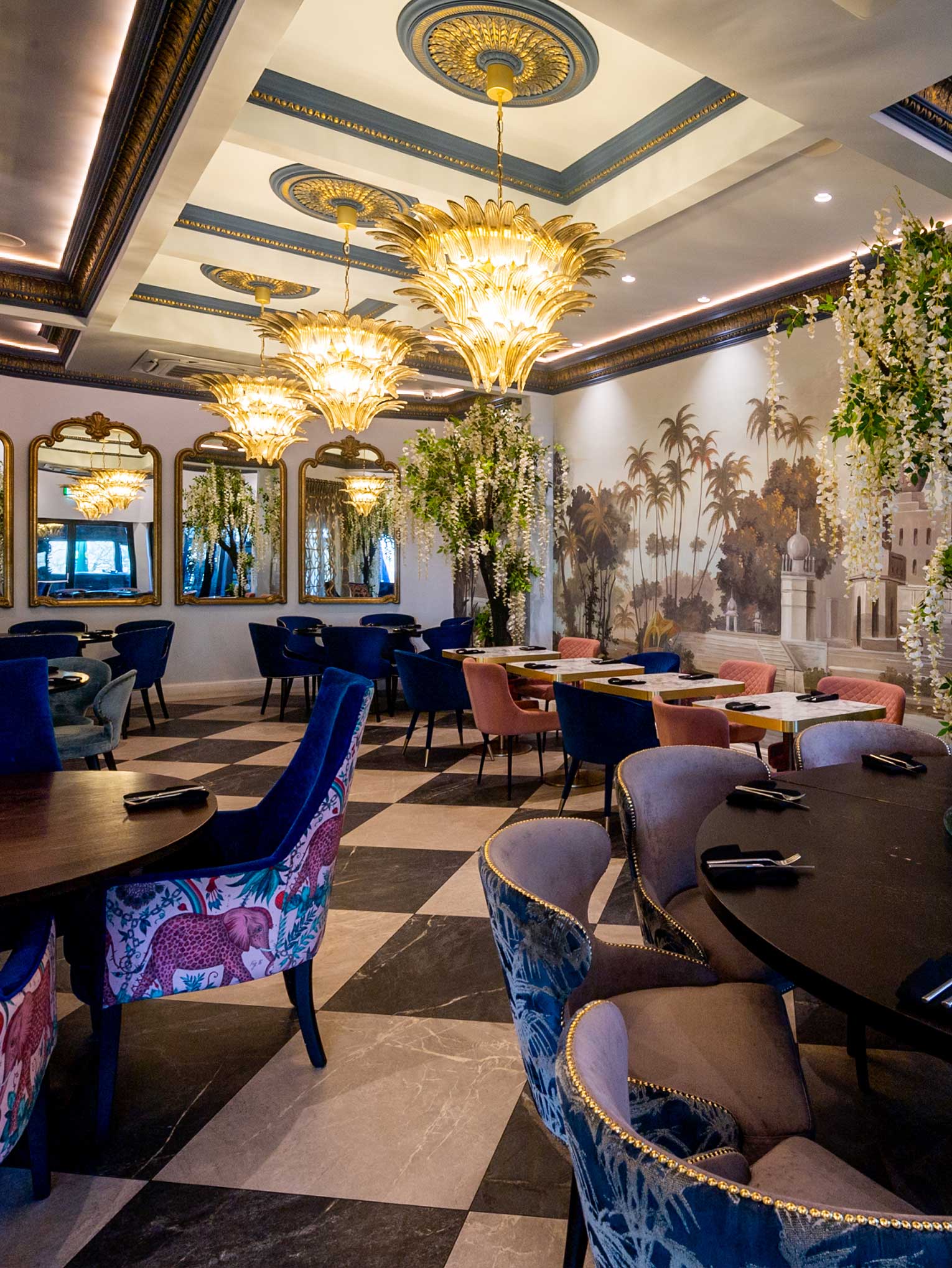 Elegant Indian restaurant interior in Stanmore with ornate chandeliers, floral upholstery, and dark wood tables. The checkered floor contrasts with a mural depicting palm trees and architecture. Mirrors line one wall, and hanging greenery adds a touch of nature. | Indian Restaurant Elstree Borehamwood