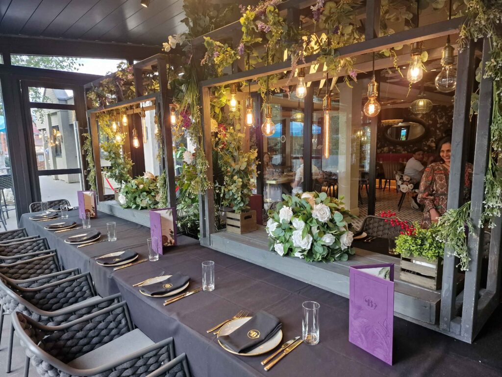 A stylish Indian Restaurant in Bushey features a sophisticated table setting with black tablecloth, gold cutlery, and black plates. Glasses are neatly placed beside each setting. Overhead hang lightbulbs amid lush greenery. A large mirror and floral arrangements decorate the space, with diners seen in the background. | Indian Restaurant Elstree Borehamwood