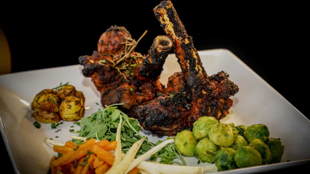 A white square plate holds grilled lamb chops garnished with herbs. Accompanying the chops are roasted potatoes, sautéed Brussels sprouts, and a mix of carrots, parsnips, and arugula, reminiscent of the vibrant flavors found at an Indian restaurant in Elstree. | Indian Restaurant Elstree Borehamwood
