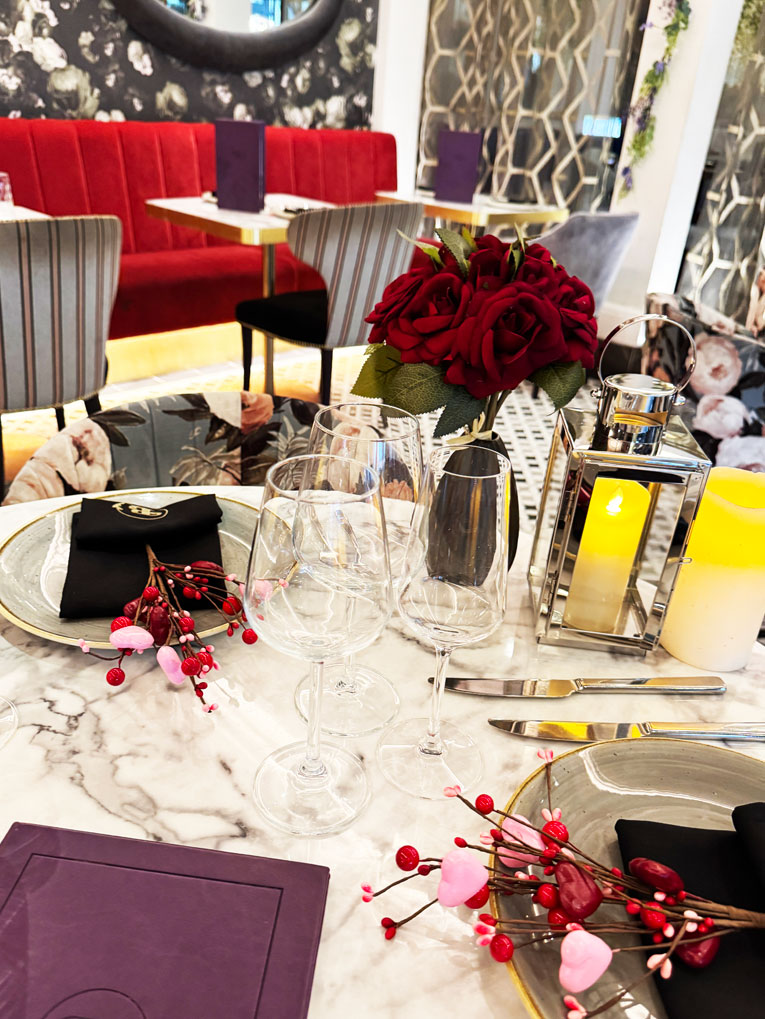 A marble table set for two with wine glasses, black napkins, and plates. Red artificial roses, a lantern with a yellow candle, and berry decor create a romantic Valentines vibe at Bhageecha, the perfect Indian restaurant near me. Plush red seating completes the scene. | Indian Restaurant Elstree Borehamwood