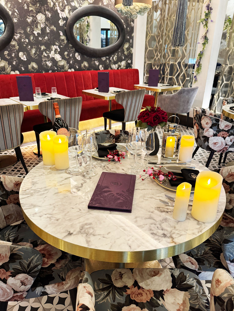 A round marble table set for dining holds LED candles, wine glasses, cutlery, a wine bottle in an ice bucket, a floral arrangement, and a purple menu—perfect for Valentines at Bhageecha, the elegant Indian restaurant near me. | Indian Restaurant Elstree Borehamwood