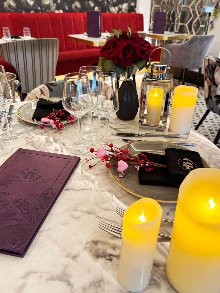 A marble table is set for dining with wine glasses, purple menus, faux candles, a vase of red roses, and festive Valentines decorations. Plush red seating and patterned black-and-white walls add charm to Bhageecha, an elegant Indian restaurant near me. | Indian Restaurant Elstree Borehamwood