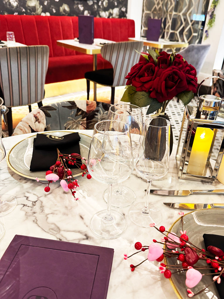 A marble restaurant table at Bhageecha is set with wine glasses, plates, black napkins, red berry and heart decorations, and a bouquet of red roses—perfect for Valentines. Red velvet seating and elegant decor complete the romantic vibe. | Indian Restaurant Elstree Borehamwood