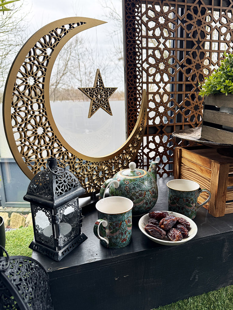 A decorative scene with a large gold crescent moon and star, a black lantern, a green teapot, two patterned mugs, and a plate of dates on a table. Ornate panels and a potted plant are in the background, with trees visible through the window. | Indian Restaurant Elstree Borehamwood