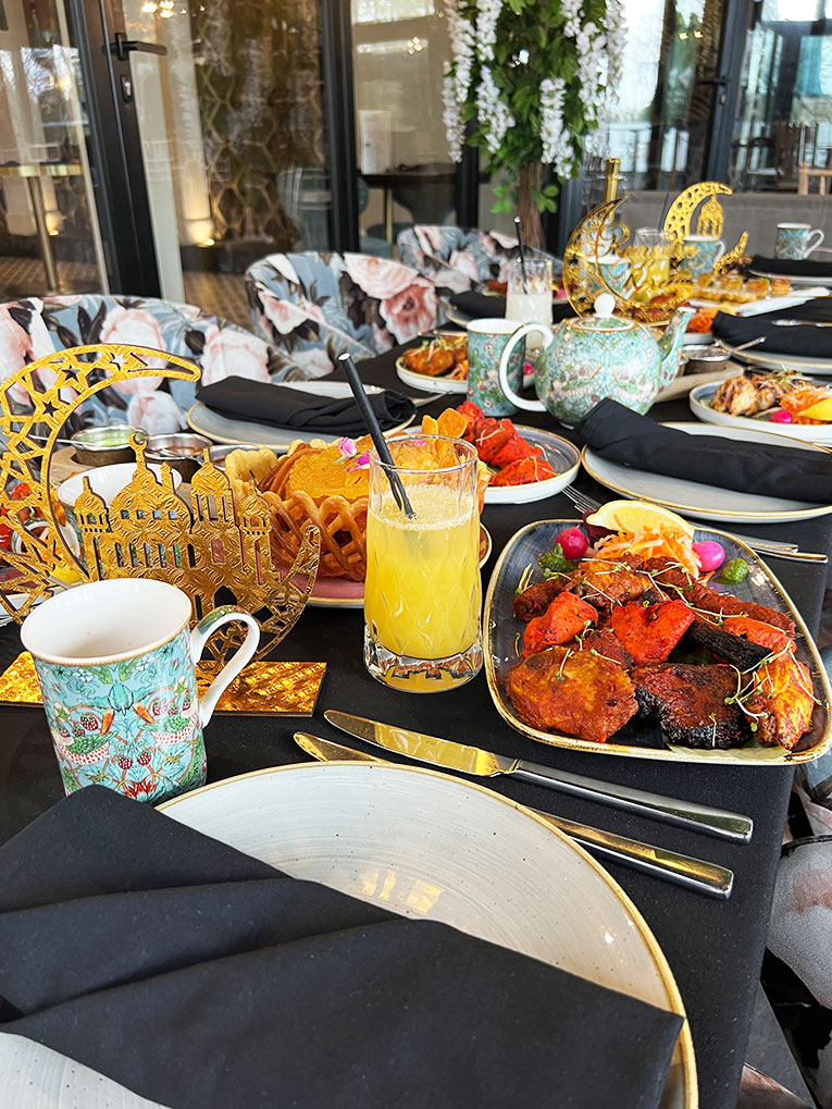 A table set for a meal with ornate plates and cups, a glass of orange juice, assorted grilled meats, fried snacks, and garnished dishes. Gold decorative pieces and a floral teapot add elegance; floral-patterned chairs and hanging flowers are in the background. | Indian Restaurant Elstree Borehamwood