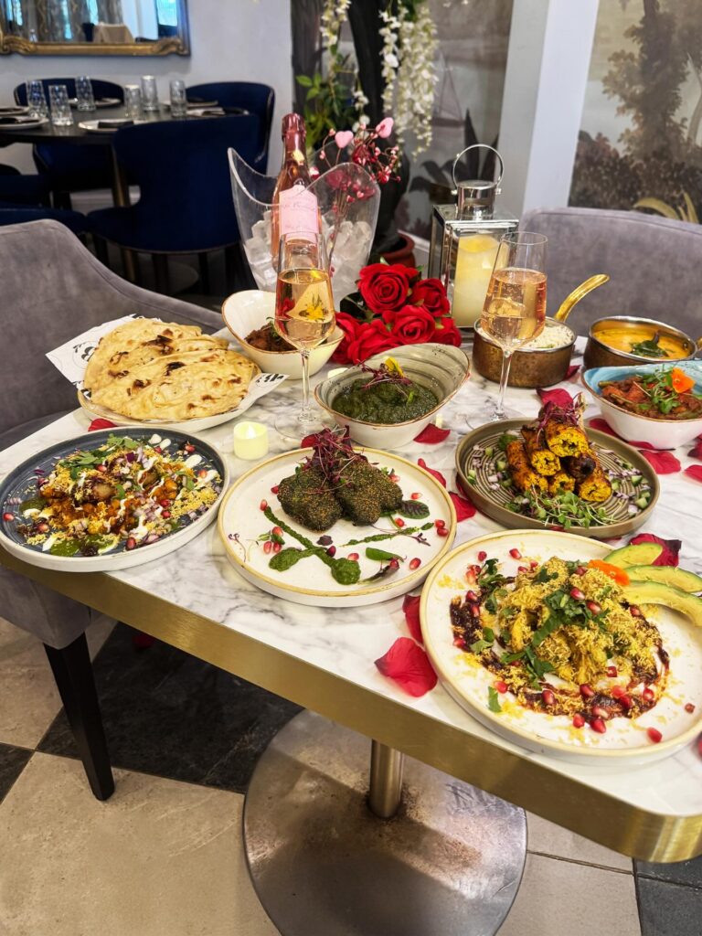 A marble table at a Modern Indian Restaurant Near Me in Church Crookham is set with assorted Indian dishes, naan, kebabs, chutneys, salads, and curries. Rosé wine, red roses, candles, and rose petals add a romantic touch to the elegant dining experience. | Indian Restaurant Elstree Borehamwood