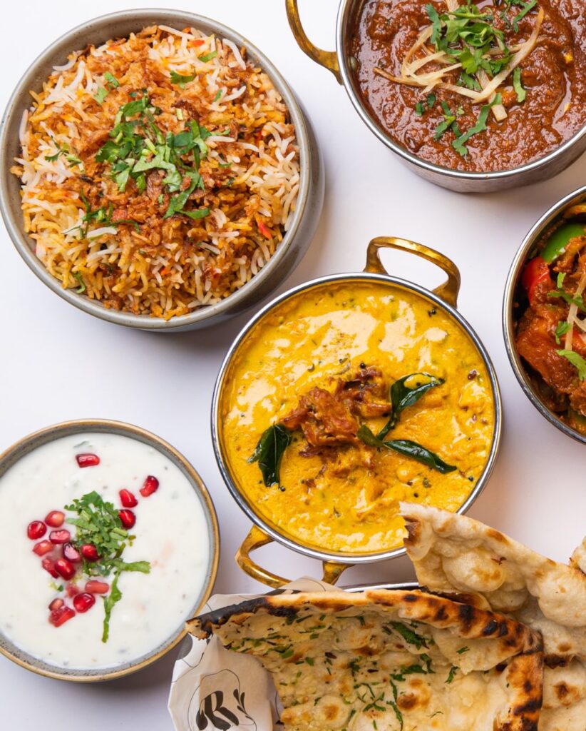 An overhead view of assorted Indian dishes at a Modern Indian Restaurant Near Me in Church Crookham: biryani rice, colourful curries, naan bread, and raita with pomegranate and herbs—all beautifully garnished with fresh coriander on a white background. | Indian Restaurant Elstree Borehamwood