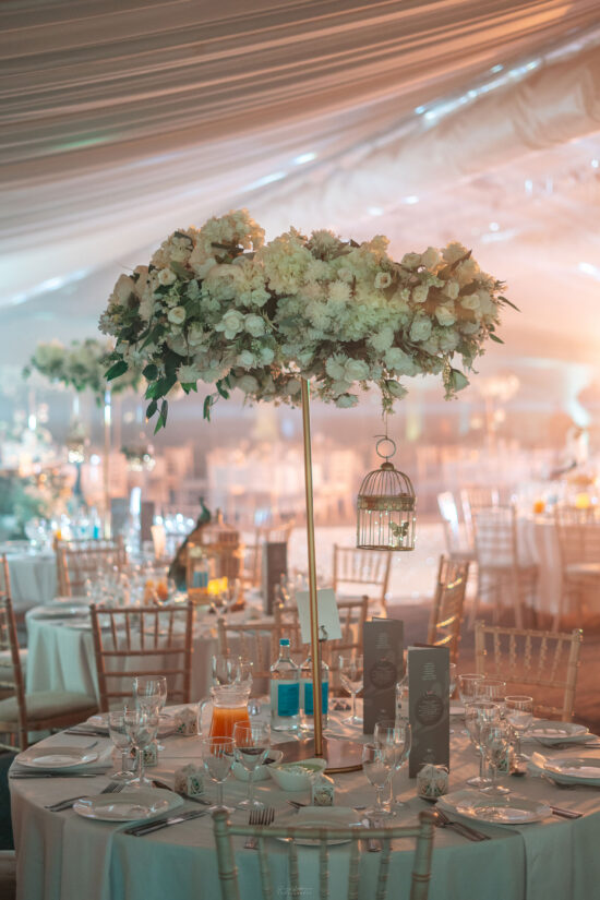 A round table set for an event at Bhageecha, a modern Indian restaurant, features white linens, elegant glassware, and menus. The tall gold centerpiece with lush white flowers and a birdcage stands out amid softly lit wooden chairs and decorated tables. | Indian Restaurant Elstree Borehamwood