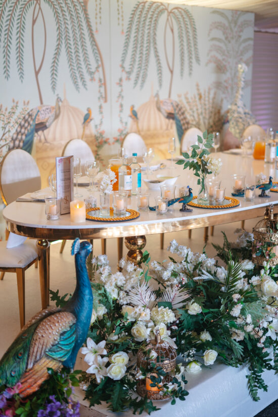 An elegant table at Bhageecha, a modern Indian restaurant, is set with glassware, candles, and floral centerpieces. Decorative peacock statues and white flowers complement the mural of palm trees and peacocks, enhancing the nature-inspired decor. | Indian Restaurant Elstree Borehamwood