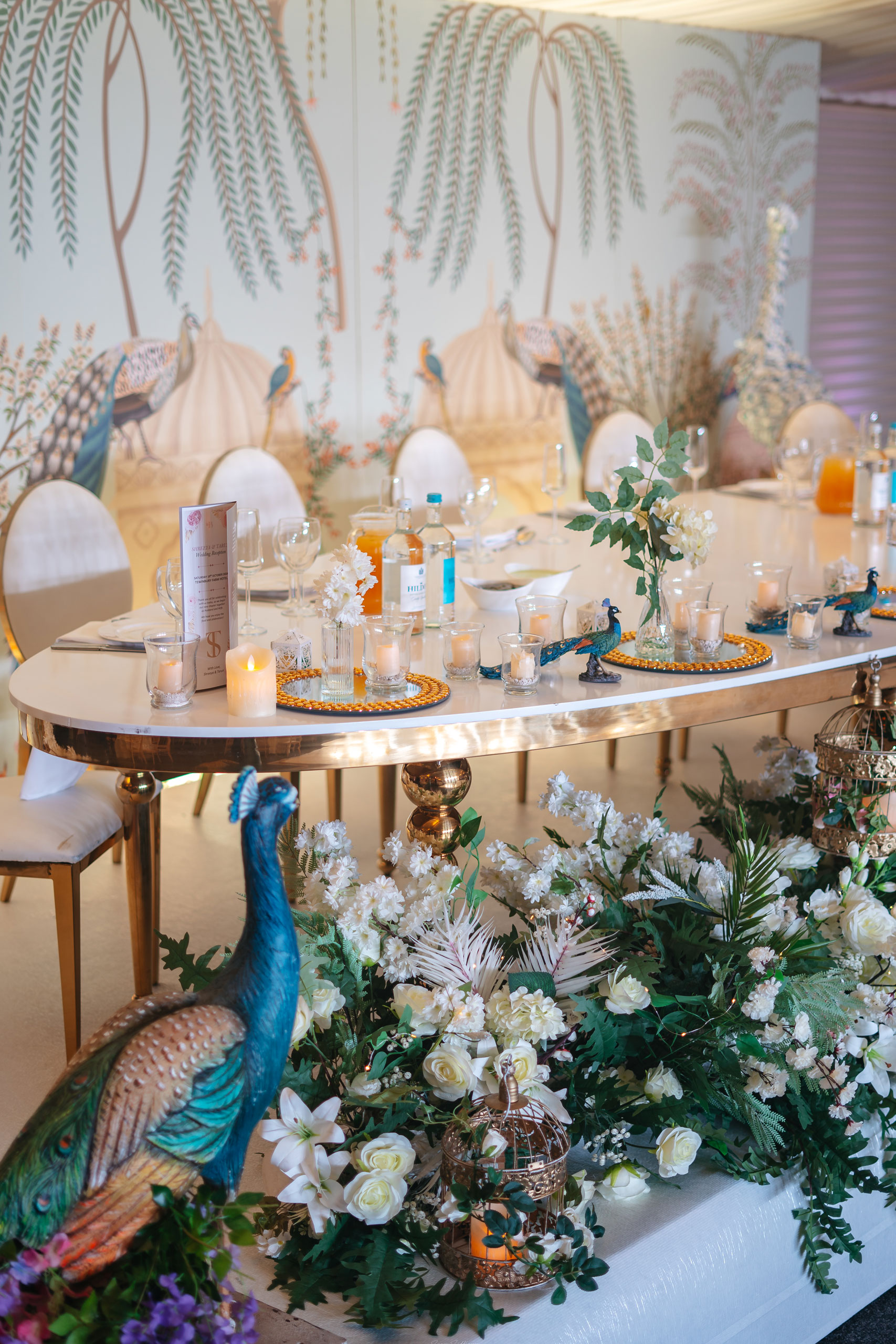 An elegant table at Bhageecha, a modern Indian restaurant, is set with glassware, candles, and floral centerpieces. Decorative peacock statues and white flowers complement the mural of palm trees and peacocks, enhancing the nature-inspired decor. | Indian Restaurant Elstree Borehamwood