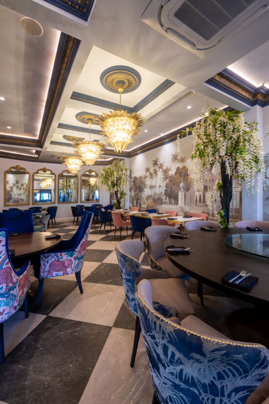 Elegant restaurant interior with ornate chandeliers, gold-trimmed ceiling, and wall mirrors. Bhageecha is a modern Indian restaurant where tables and velvet chairs sit among lush green plants and painted palm murals in a bright, stylish space. | Indian Restaurant Elstree Borehamwood