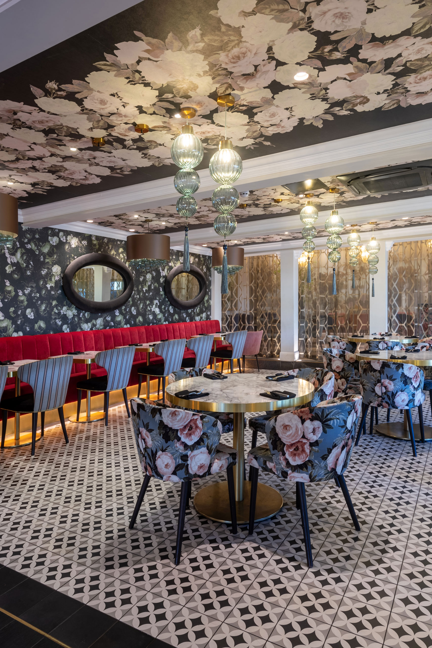 A stylish modern Indian restaurant with floral-patterned chairs, a matching floral ceiling, patterned floor tiles, and pink cushioned bench seating. Round mirrors hang on a dark floral wall as glass pendant lights add elegance to this luxurious bhageecha. | Indian Restaurant Elstree Borehamwood