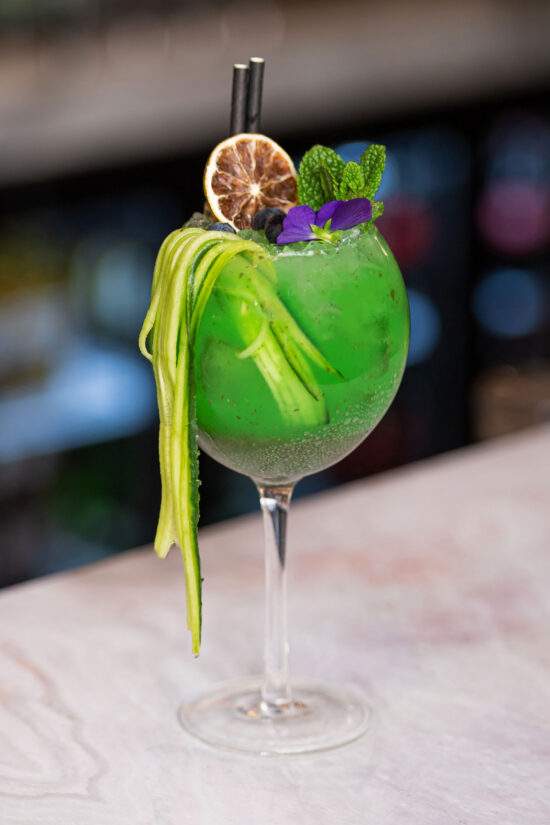 A vibrant green cocktail in a stemmed glass is garnished with cucumber ribbons, dried lemon, mint sprigs, and edible flowers at Bhageecha—an inviting spot if you're searching for an Indian restaurant near me or the cozy Bhageecha Elstree Rooms. | Indian Restaurant Elstree Borehamwood