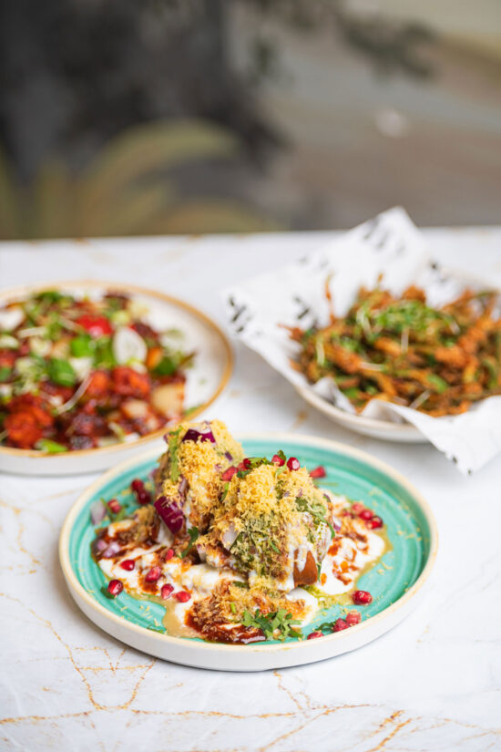 A vibrant plate of Indian chaat from Bhageecha Elstree Rooms sits in focus, topped with yogurt, chutneys, sev, cilantro, and pomegranate seeds. A leafy salad and crispy fried greens complete the scene at this inviting Indian restaurant near me. | Indian Restaurant Elstree Borehamwood