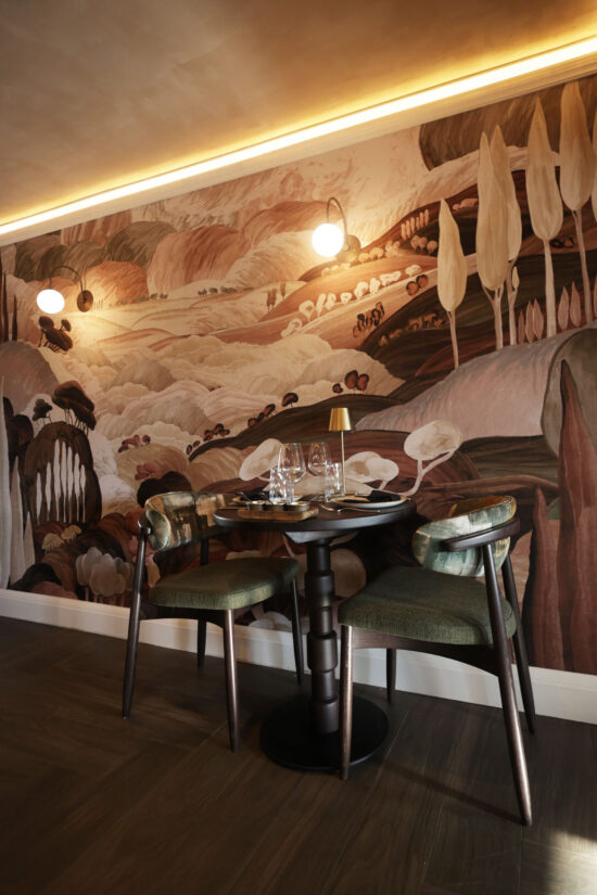 A round dining table set for two with green cushioned chairs sits against a mural wall featuring abstract, earthy-toned landscapes and trees. Warm lighting from wall sconces creates a cozy, elegant vibe at this modern Indian restaurant, Bhageecha. | Indian Restaurant Elstree Borehamwood