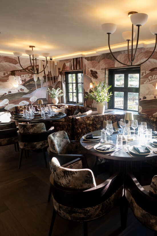 Elegant modern Indian restaurant interior with round tables, upholstered chairs, and floor-to-ceiling wall art inspired by abstract nature scenes. Large windows and chandeliers create a warm, inviting ambiance at Bhageecha—your Indian restaurant near me. | Indian Restaurant Elstree Borehamwood