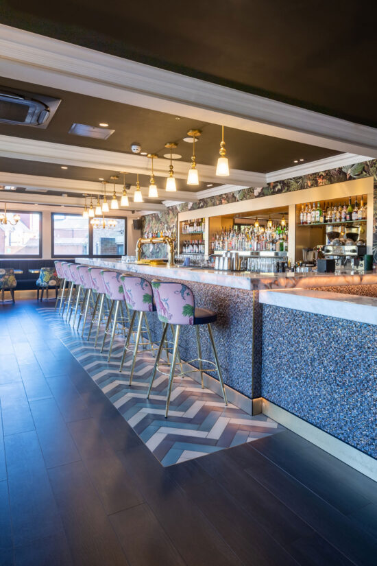 Modern bar interior at Bhageecha Elstree Rooms with a patterned tile counter, gold pendant lights, and pastel pink bar stools featuring palm leaf designs—perfect for those searching for a stylish Indian restaurant near me. | Indian Restaurant Elstree Borehamwood