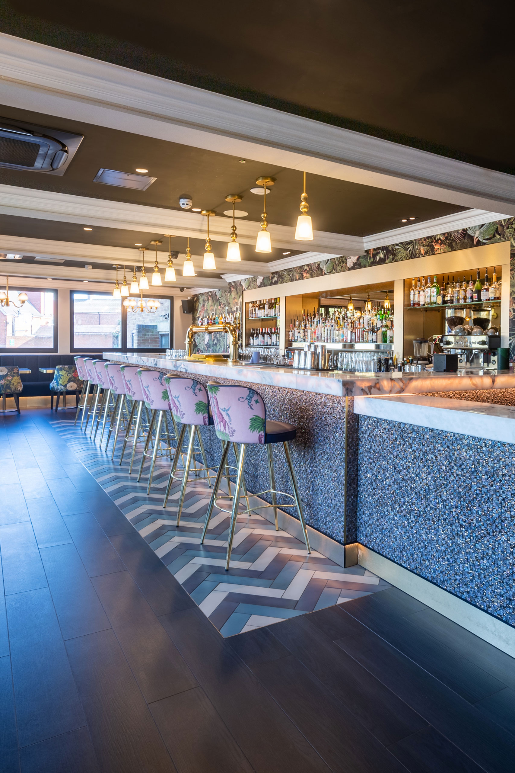 Modern bar interior at Bhageecha Elstree Rooms with a patterned tile counter, gold pendant lights, and pastel pink bar stools featuring palm leaf designs—perfect for those searching for a stylish Indian restaurant near me. | Indian Restaurant Elstree Borehamwood