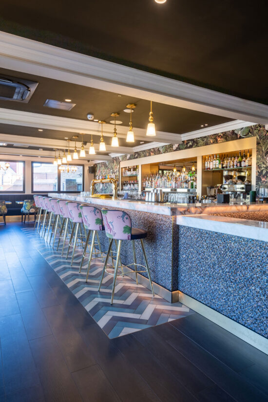 A stylish modern Indian restaurant with a patterned tile floor, marble countertop, and gold accents. Pink bar stools with floral designs line the counter, while shelves of spirits and pendant lights shine under large, natural light-filled windows. | Indian Restaurant Elstree Borehamwood