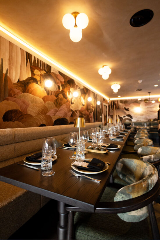 A modern Indian restaurant with elegant table settings, including wine glasses, black napkins, and gold plates. Plush seating, artistic wall murals, and warm ambient lighting from globe fixtures create a stylish bhageecha-inspired dining space. | Indian Restaurant Elstree Borehamwood