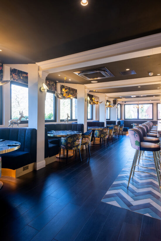 A modern, stylish Indian restaurant near me with dark wood floors, blue velvet booths, floral-patterned chairs, and a long bar with pastel stools. Large windows let in natural light and decorative lighting fixtures hang from the ceiling. | Indian Restaurant Elstree Borehamwood