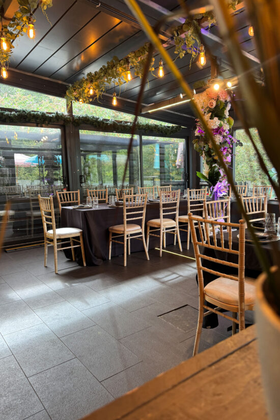 A modern indoor dining area at Bhageecha Elstree Rooms features wood chairs around tables covered in dark cloths. Overhead, string lights and green foliage are draped decoratively, evoking a serene bhageecha vibe with out-of-focus plant leaves in the foreground. | Indian Restaurant Elstree Borehamwood