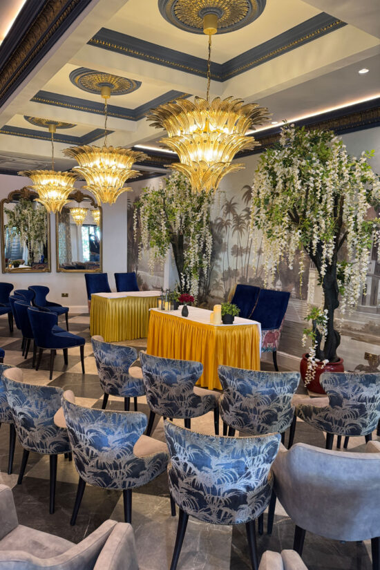 Elegant indoor event setup with blue and gray patterned chairs, reminiscent of a modern Indian restaurant. Ornate chandeliers glow above, while cascading white flowers and a decorative mural create an ambiance that rivals Bhageecha, the finest indian restaurant near me. | Indian Restaurant Elstree Borehamwood