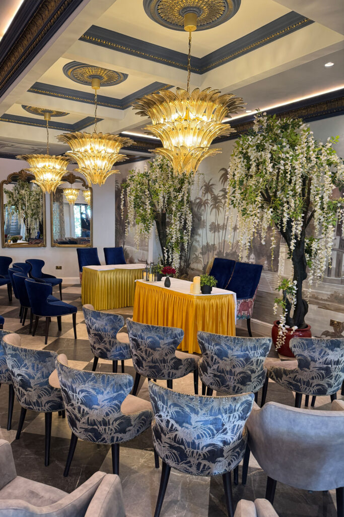 Elegant indoor event setup with blue and gray patterned chairs, reminiscent of a modern Indian restaurant. Ornate chandeliers glow above, while cascading white flowers and a decorative mural create an ambiance that rivals Bhageecha, the finest indian restaurant near me. | Indian Restaurant Elstree Borehamwood