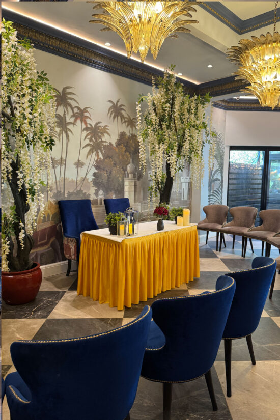 A formal room at Bhageecha, a modern Indian restaurant, features a table draped in yellow cloth, flanked by blue velvet chairs. Tall artificial trees with white flowers add charm, while ornate chandeliers and a palm tree mural complete the look. | Indian Restaurant Elstree Borehamwood