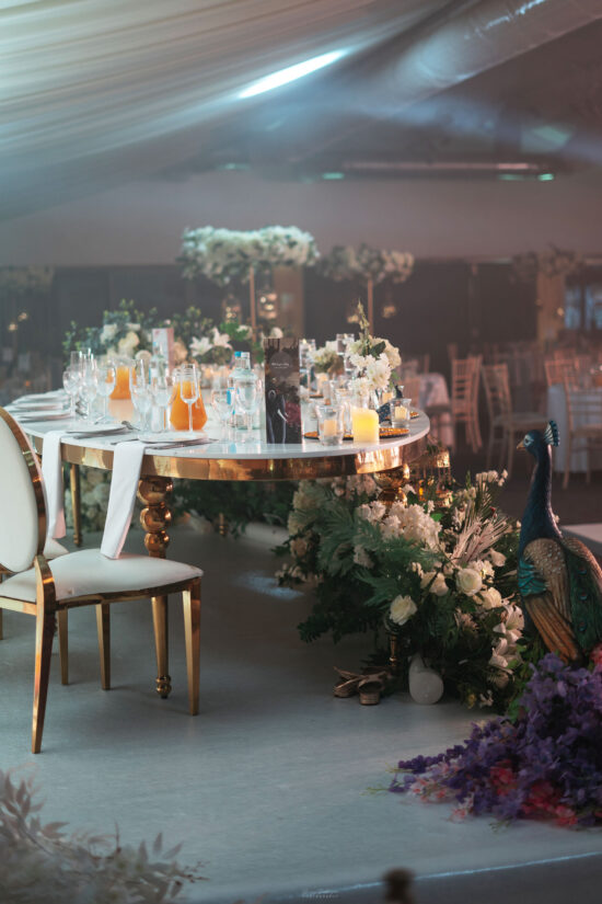 A lavishly decorated round table with a gold base is set for an event at Weddings at Bhageecha, featuring glassware, orange juice, candles, and floral arrangements. White chairs surround the table and a decorative peacock stands beside lush flowers. | Indian Restaurant Elstree Borehamwood