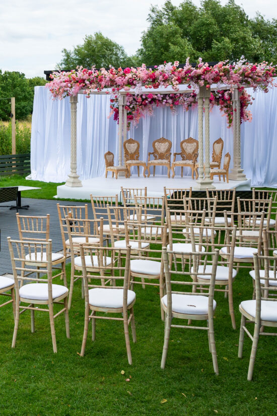 Rows of empty wooden chairs with white cushions face a decorated wedding mandap at Bhageecha, draped in white fabric and a canopy of pink, purple, and white flowers—perfect for outdoor weddings at Bhageecha, not just your favorite Indian restaurant near me. | Indian Restaurant Elstree Borehamwood