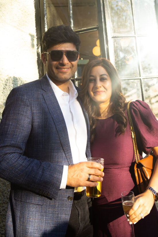 A man in a blue blazer and sunglasses stands next to a woman in a purple dress. Both are smiling and holding drinks, with sunlight streaming in at Bhageecha, a modern Indian restaurant, near a window with reflections and a textured wall. | Indian Restaurant Elstree Borehamwood