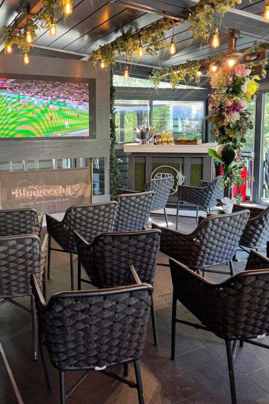A modern indoor seating area at Bhageecha features woven black chairs facing a TV showing The World Cup At Bhageecha Elstree. Warm fairy lights and greenery hang above, while the bar counter glows with flowers and bottles under large windows. | Indian Restaurant Elstree Borehamwood