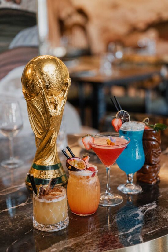 A gold FIFA World Cup trophy stands on a marble table at Bhageecha beside four colourful cocktails garnished with fresh fruit and straws, set in an inviting Indian restaurant near me with a blurred dining area in the background. | Indian Restaurant Elstree Borehamwood