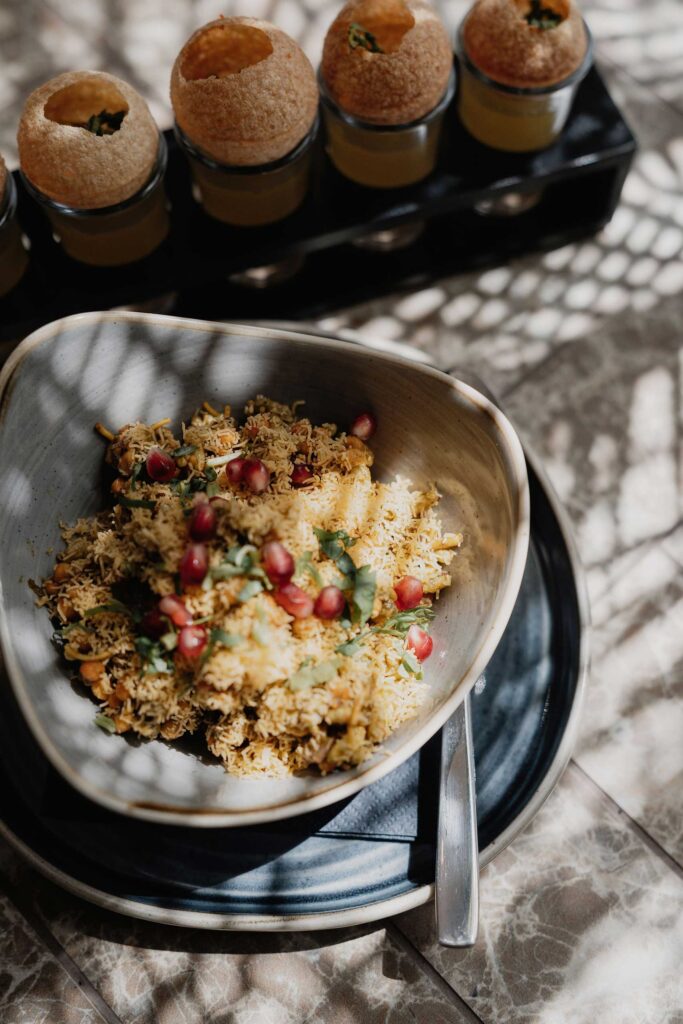 A bowl of yellow rice garnished with pomegranate seeds and herbs sits on a dark plate with a spoon. In the background, round crispy puris are served atop filled shot glasses. Sunlight casts patterned shadows across the table. | Indian Restaurant Elstree Borehamwood