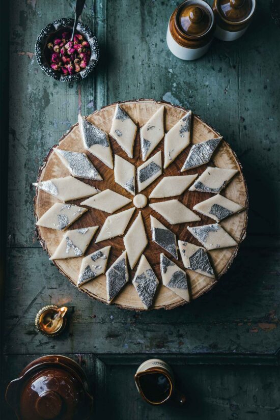 A round wooden board with diamond-shaped Indian sweets, some topped with silver leaf, evokes the authentic flavours you’d find at Bhageecha Hampshire Private Hire. Nearby are a small dish with dried rosebuds, two ceramic jars, a lit oil lamp, and a teapot on a rustic green table, setting the scene reminiscent of an elegant Indian restaurant near me. | Indian Restaurant Elstree Borehamwood