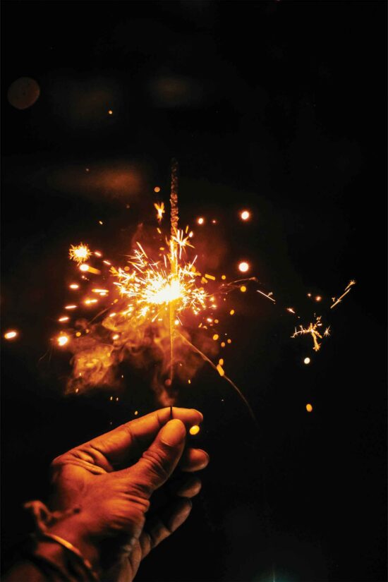 A hand holds a lit sparkler in the dark, with bright orange and yellow sparks radiating outward, creating a glowing, festive atmosphere against a black background—reminiscent of celebrations at Bhageecha Hampshire Private Hire. Wisps of smoke swirl around the hand and the sparkler, echoing the vibrant spirit found at your favourite Indian restaurant near me. | Indian Restaurant Elstree Borehamwood