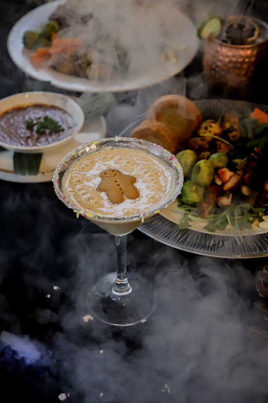A cocktail in a sugar-rimmed martini glass is topped with a gingerbread biscuit. Around it are plates of food and a copper mug, all surrounded by swirling smoke, creating a dramatic effect—perfect for celebrating Christmas at Hampshire in the cosy ambience of bhageecha, the finest Indian restaurant near me. | Indian Restaurant Elstree Borehamwood