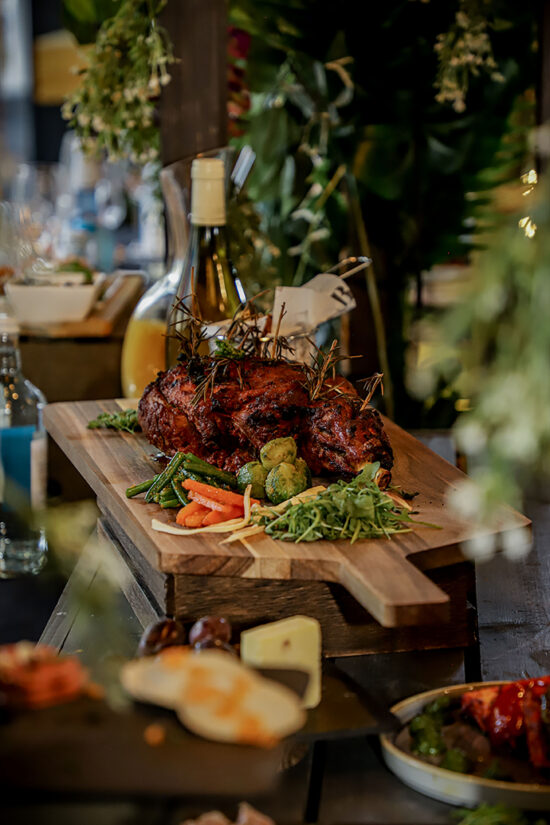 A roast meat dish garnished with herbs sits on a wooden board, surrounded by roasted vegetables, microgreens, and a small bunch of carrots. A bottle of white wine and leafy greenery are in the background, enhancing the rustic table setting—perfect for celebrating Christmas at Hampshire or enjoying festive flavours inspired by Bhageecha. | Indian Restaurant Elstree Borehamwood