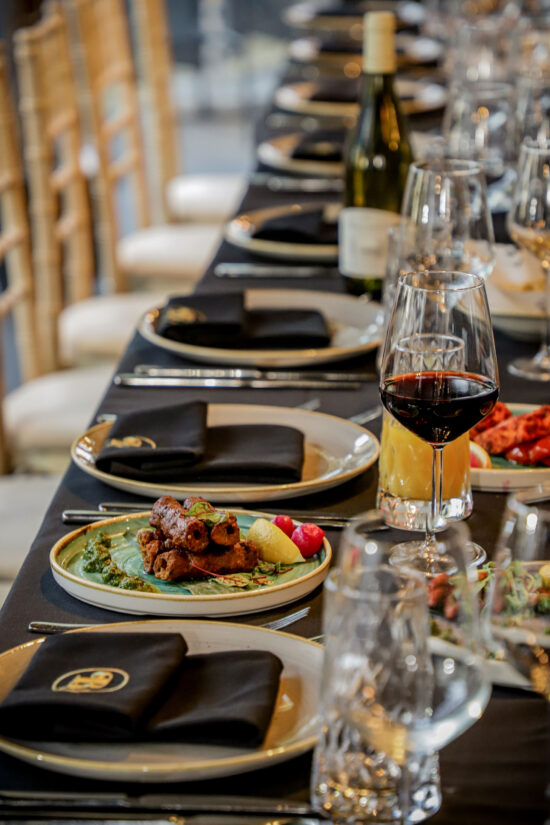 A formal dining table set with black serviettes, plates of gourmet food, wine glasses with red and white wine, and a bottle of white wine. Gold chairs line the table, with intricate food presentation and elegant table settings in a restaurant or event space—perfect for celebrating Christmas in Hampshire or enjoying a luxurious evening at Bhageecha, an exceptional Indian restaurant near me. | Indian Restaurant Elstree Borehamwood
